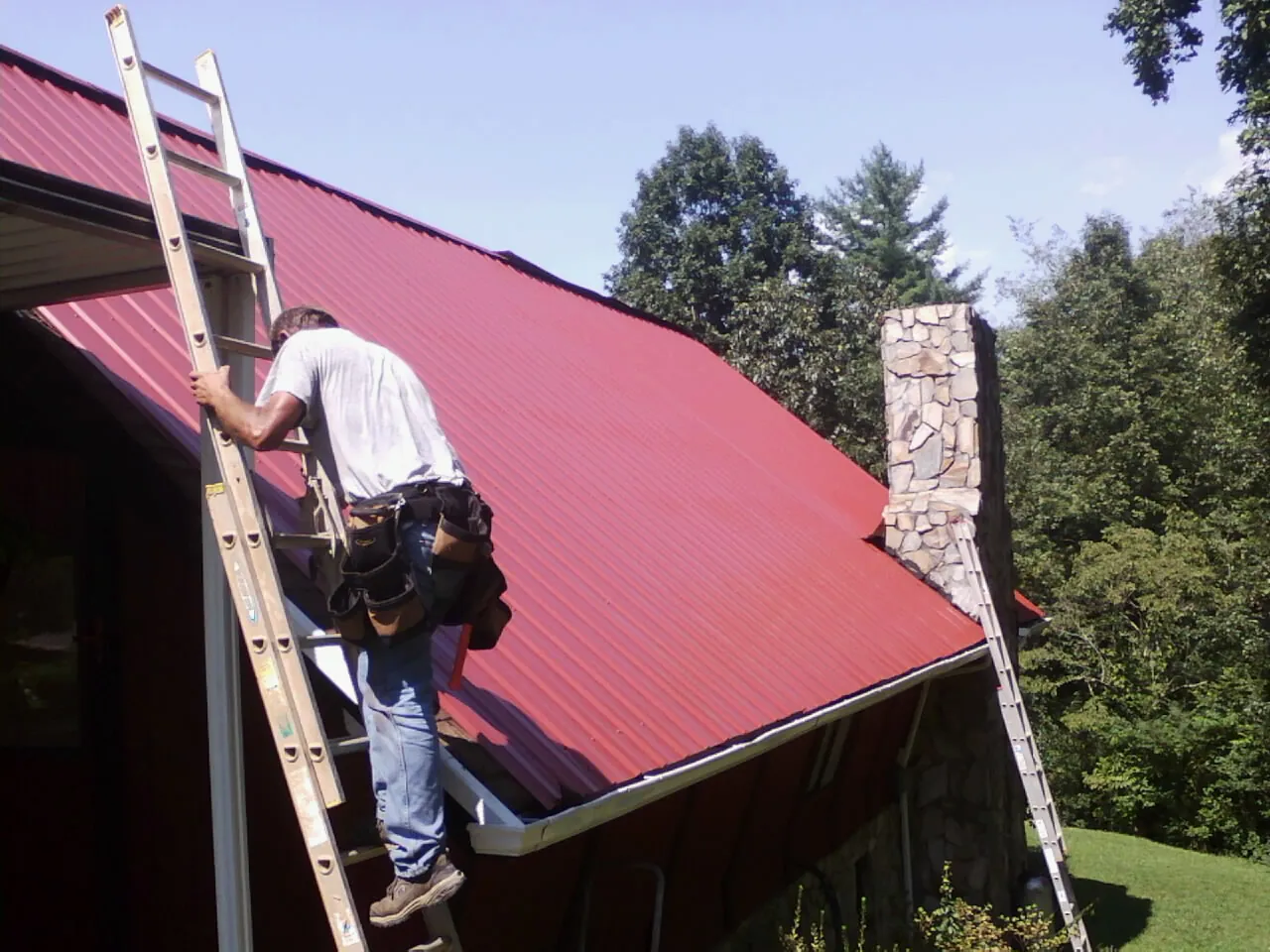 Roofer on a ladder beside a red metal roof and stone chimney — Soffit & Fascia Repair by Roofing Brothers in Yale